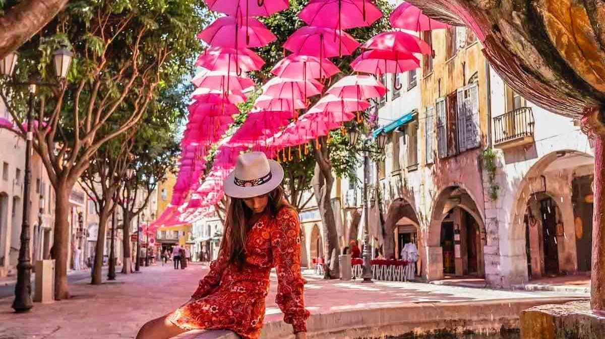 Charming Grasse street scene with pink umbrellas suspended above a sun-dappled pedestrian lane lined with Provençal buildings