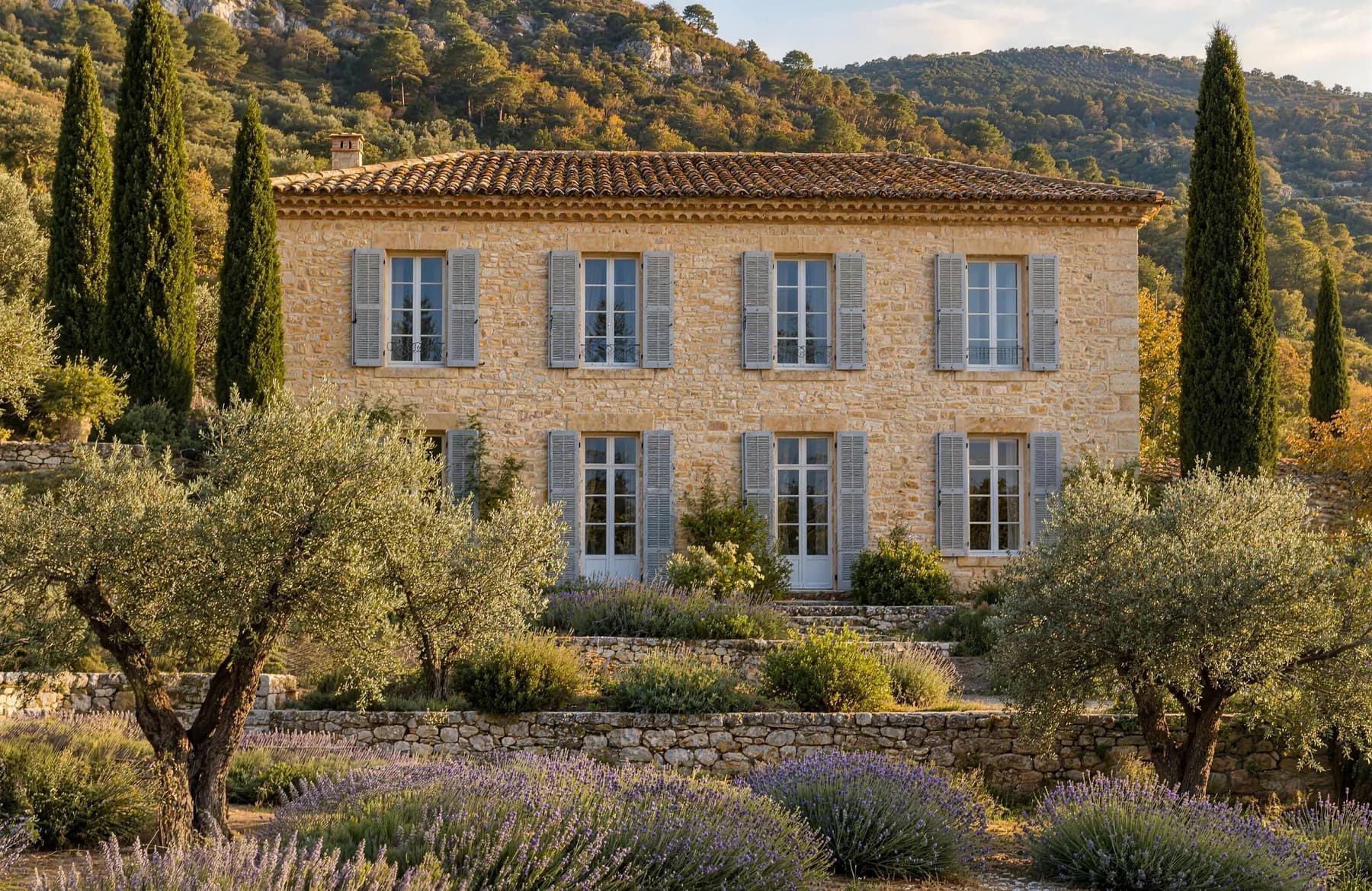Stone bastide with white shutters above terraced olive groves, lavender and dry-stone walls in late afternoon light