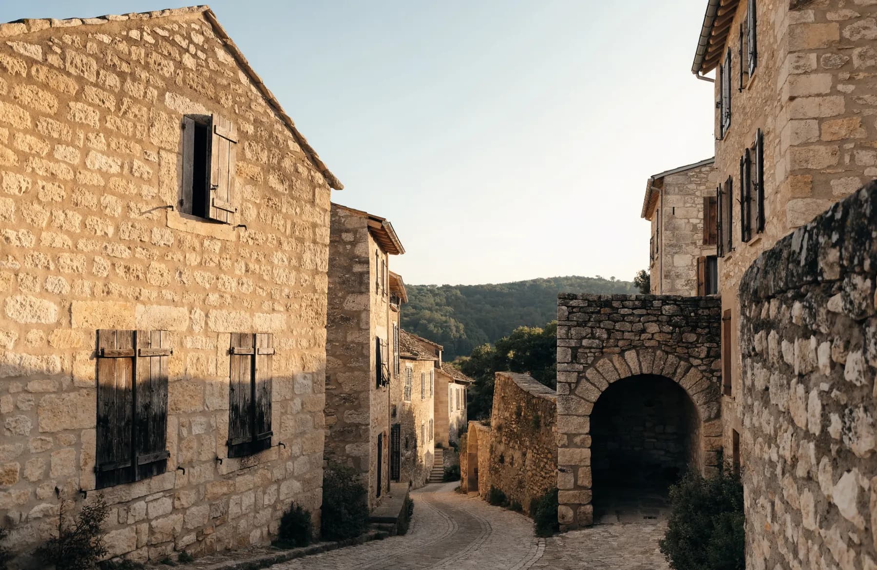 Quiet old Provençal hilltop village lane at golden hour with warm stone houses, weathered shutters and a stone archway