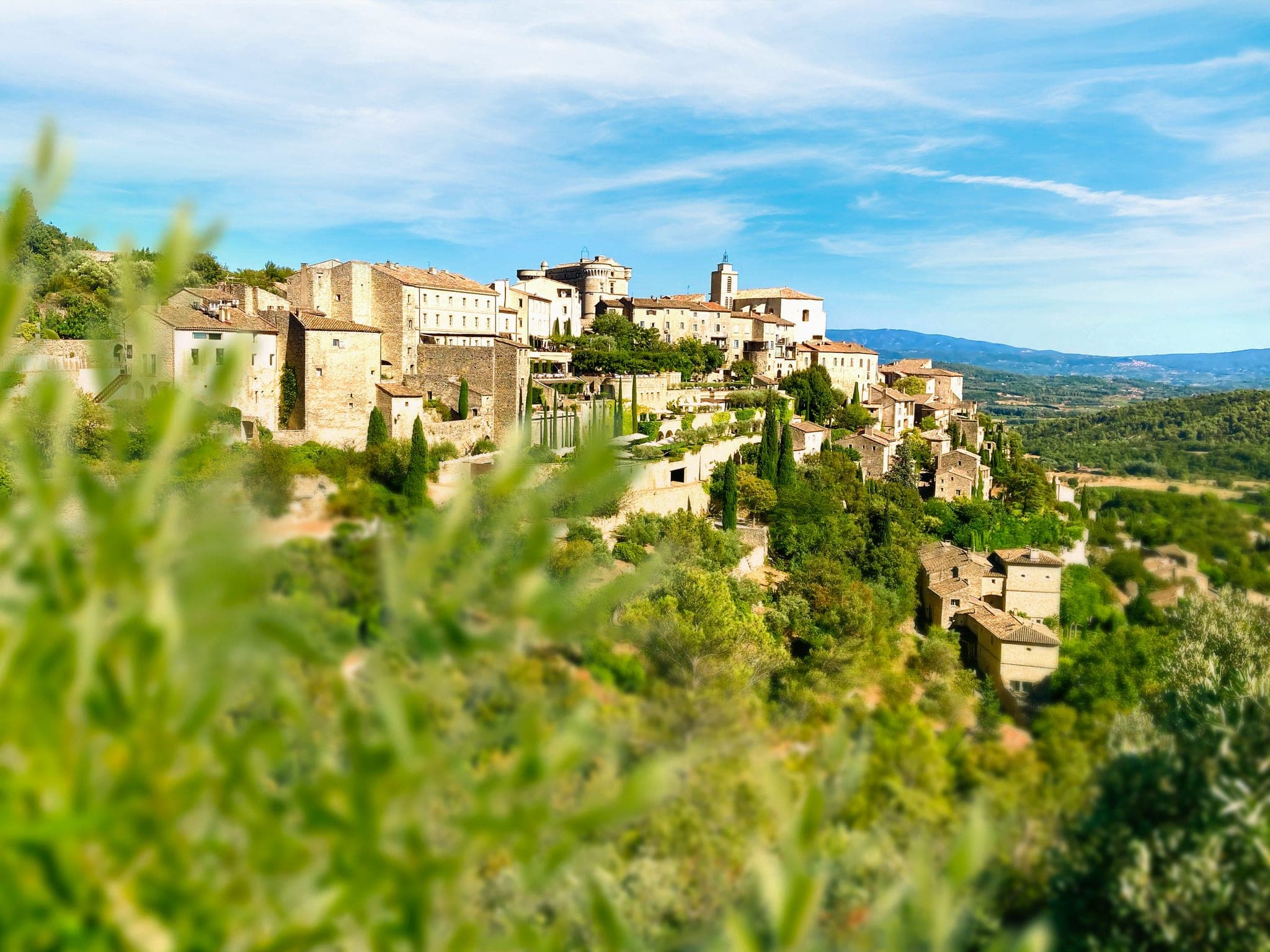 Terrasse en pierre tachetée de soleil dans l'arrière-pays provençal à l'heure de l'apéro, avec oliviers et piscine paisible en arrière-plan.