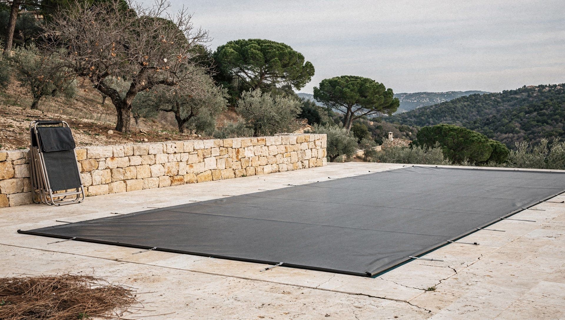 A swimming pool sealed under a dark winter cover on a travertine terrace above a wooded valley near Le Rouret in the Riviera hinterland, under overcast winter light