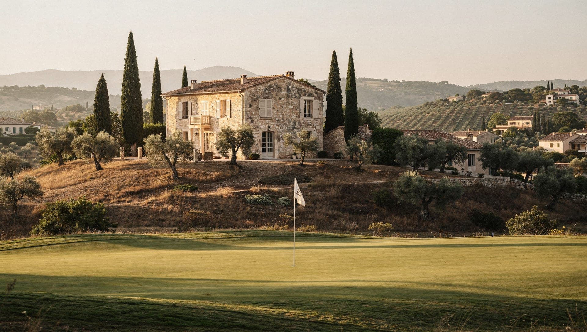 Fairway and umbrella pines on a hinterland golf course above Cannes at golden hour, stone villas beyond.