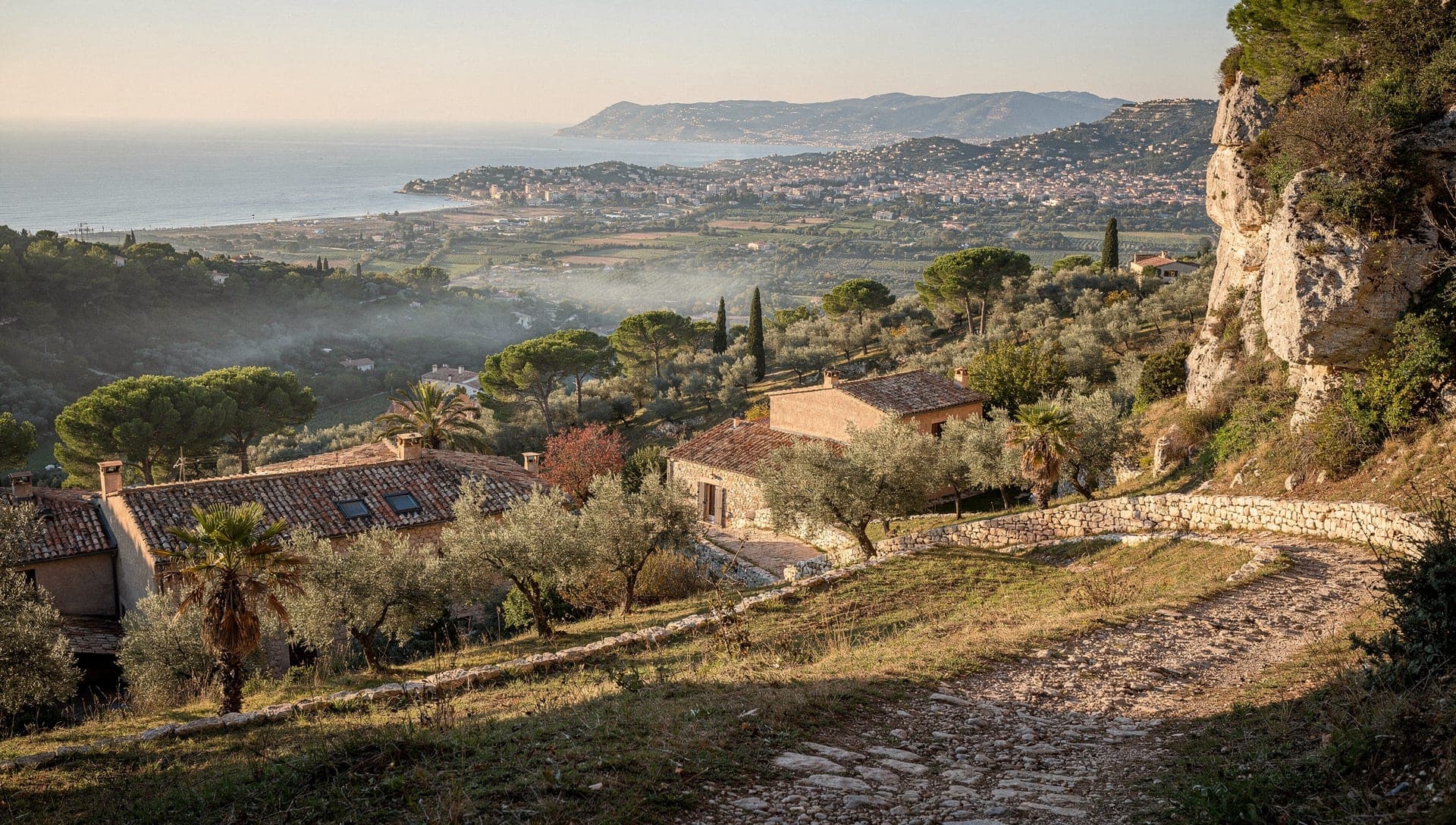 Early morning view from a stony hillside path above Chateauneuf-de-Grasse over olive terraces, stone houses and the Opio plain toward the Bay of Cannes