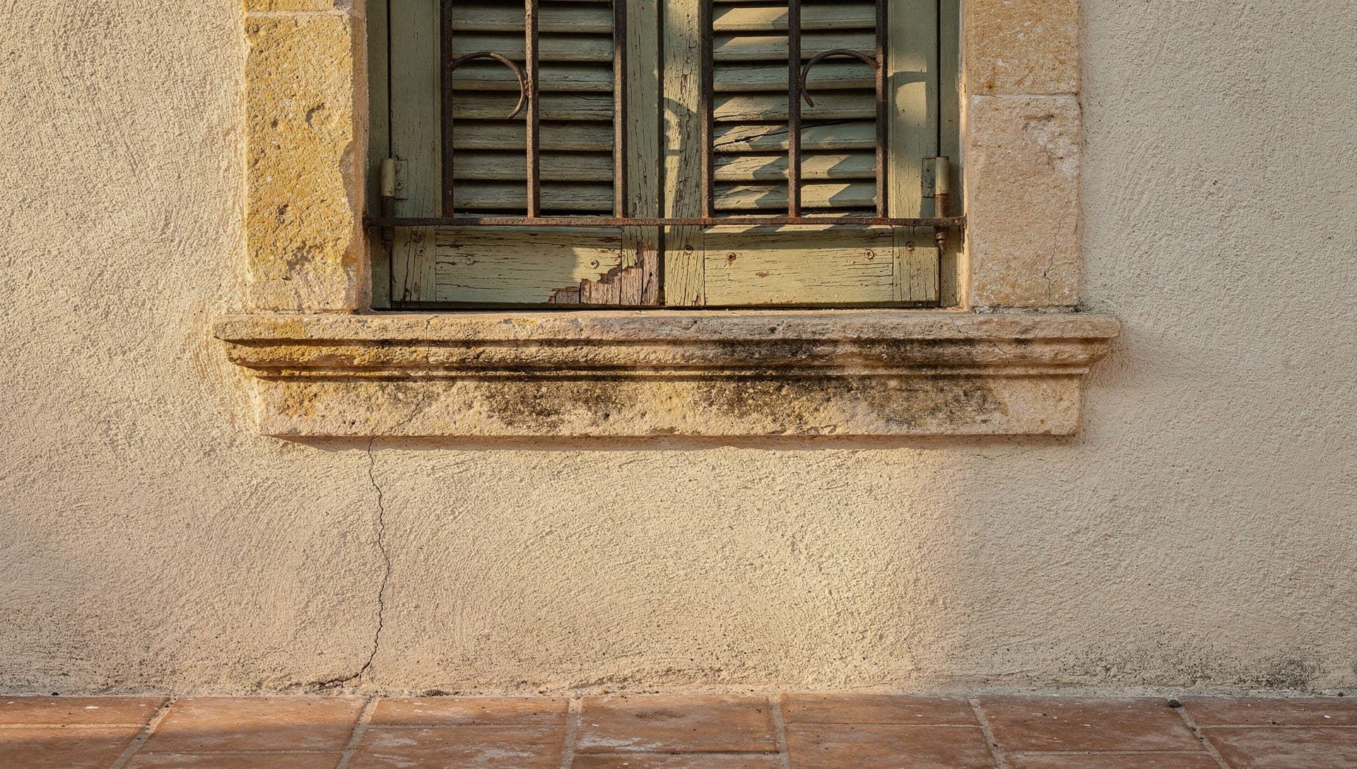 Sage-green timber shutter, limestone sill and a hairline crack in the lime render of a Provencal village house in the Riviera hinterland at early morning